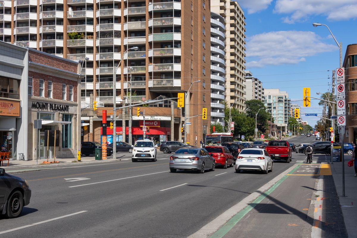 Shops, apartment buildings and traffic on Eglinton Avenue East at Mount Pleasant Road, with the Mount Pleasant station building on the corner.